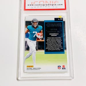 May include: A graded Panini Donruss football card featuring Brian Thomas Jr. of the Jacksonville Jaguars. The card is encased in a clear protective holder with the Iconic Grading Co. logo at the top. The player is wearing a teal jersey with the number 7.
