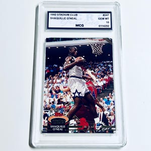 May include: A 1992 Stadium Club Shaquille O'Neal trading card in a protective case. The card shows Shaquille O'Neal playing basketball. The card is graded GEM MT 10 by MCG.