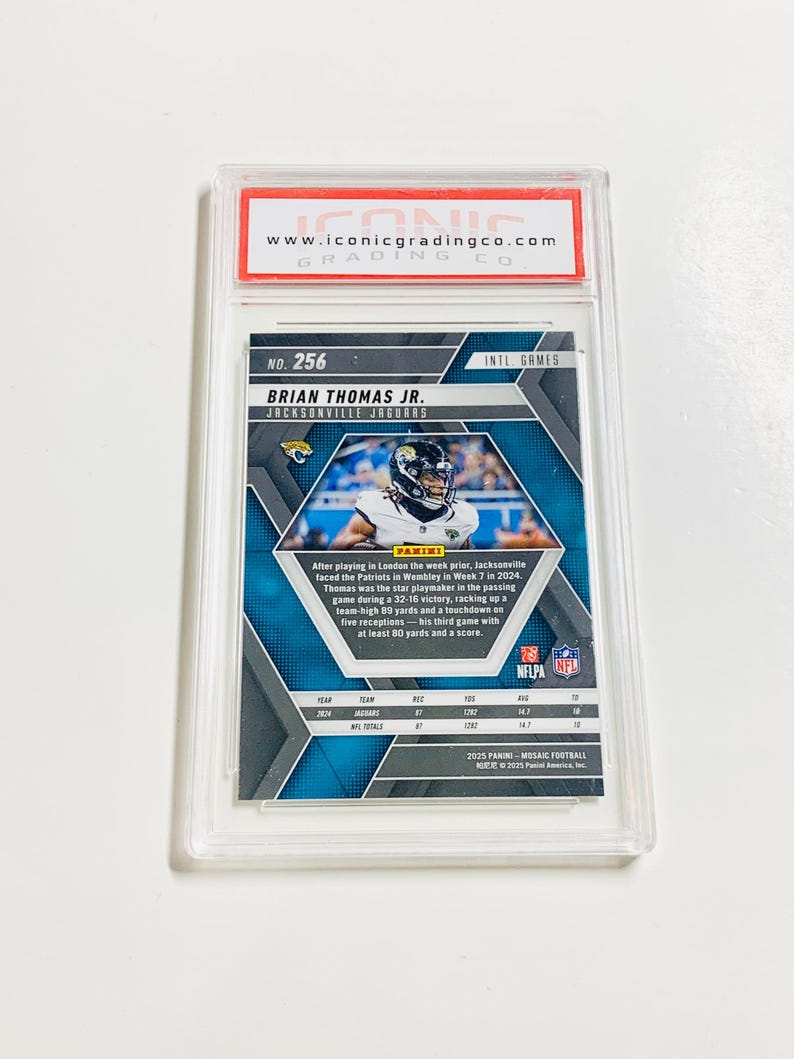 May include: A graded Brian Thomas Jr. Jacksonville Jaguars football card, encased in a clear protective holder. The card features a mosaic design and includes text about the player's performance. The top of the holder has a red and white label with the website address.