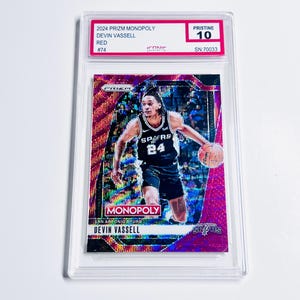 Op de afbeelding: Een rode en iriserende 2024 Prizm Monopoly trading card van Devin Vassell in een beschermhoes. De kaart toont een foto van Vassell in een Spurs-uniform. De kaart is beoordeeld met Pristine 10.