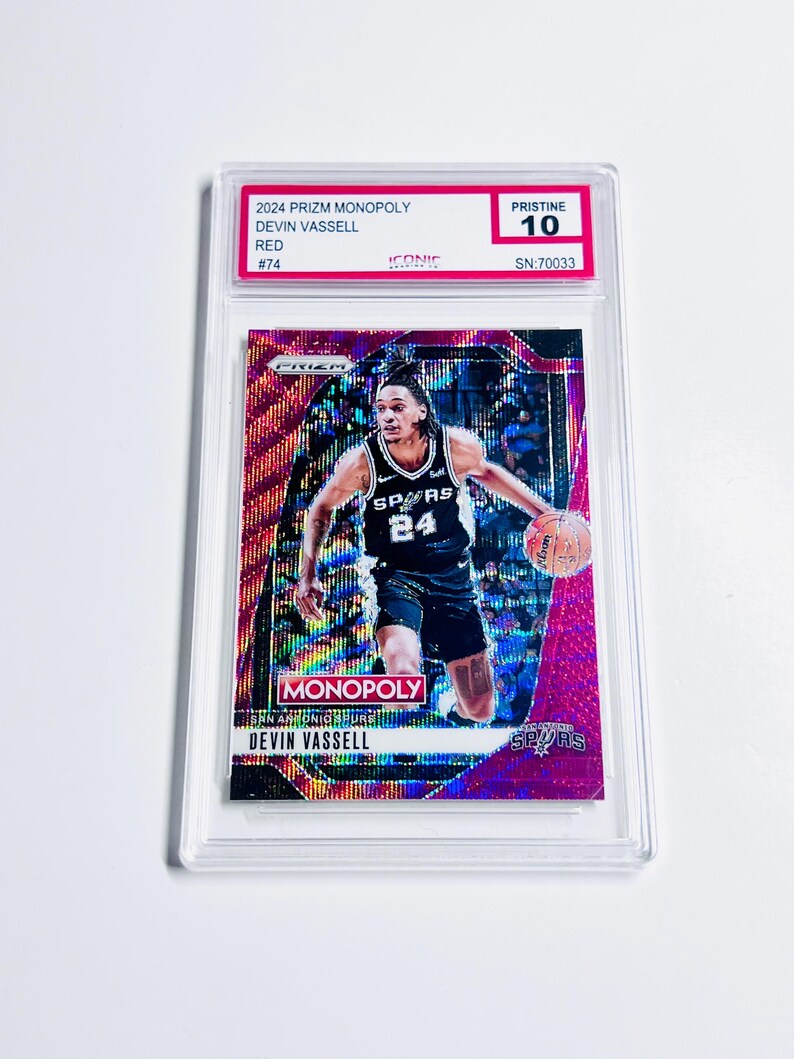 K&ouml;nnte beinhalten: Eine rote und schillernde 2024 Prizm Monopoly Trading Card von Devin Vassell in einer Schutzh&uuml;lle. Die Karte zeigt ein Foto von Vassell in einem Spurs-Trikot. Die Karte ist mit Pristine 10 bewertet.