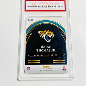 May include: A graded trading card featuring Brian Thomas Jr. of the Jacksonville Jaguars. The card is encased in a clear plastic holder with the "ICONIC GRADING CO" logo at the top. The card has a gold, black, and teal design with the Panini logo.
