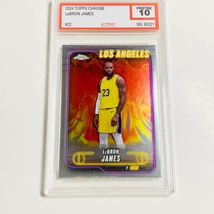 May include: A LeBron James basketball card from the 2024 Topps Chrome series, graded Pristine 10. The card features James in a yellow jersey with the number 23, set against a fiery background. The card is encased in a protective holder.