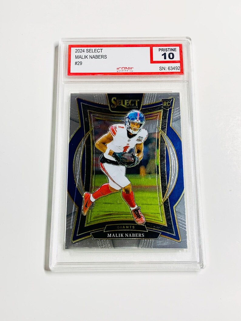 Puede incluir: Una tarjeta coleccionable de f&uacute;tbol con Malik Nabers con el uniforme de los Giants. La tarjeta est&aacute; calificada como Pristine 10 y est&aacute; dentro de un soporte protector. Se puede leer el texto "2024 SELECT MALIK NABERS #29" y "GIANTS MALIK NABERS".