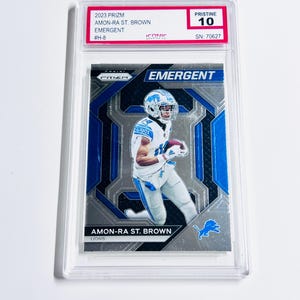 May include: A graded Amon-Ra St. Brown football card from the 2023 Prizm set. The card features a player in a Detroit Lions uniform, with the word "EMERGENT" prominently displayed. The card is graded Pristine 10 and encased in a protective holder.