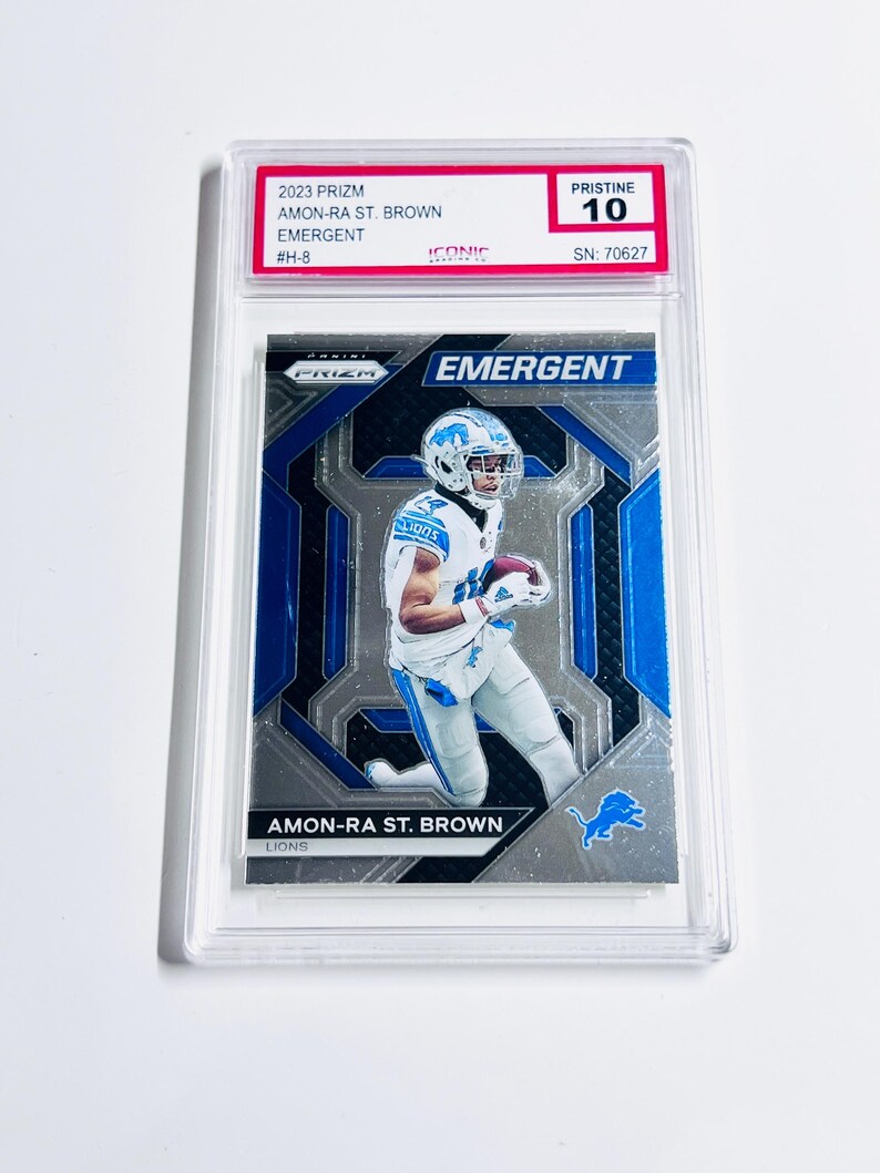 May include: A graded Amon-Ra St. Brown football card from the 2023 Prizm set. The card features a player in a Detroit Lions uniform, with the word "EMERGENT" prominently displayed. The card is graded Pristine 10 and encased in a protective holder.