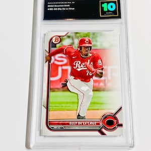 May include: A graded baseball card featuring Elly De La Cruz in a red Cincinnati Reds uniform. The card is encased in a clear plastic holder with a grade of 10. Text includes "2022 Bowman Draft" and "Elly De La Cruz".
