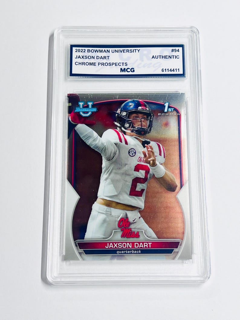 K&ouml;nnte beinhalten: Eine 2022 Bowman University Chrome Prospects Sammelkarte von Jaxson Dart, von MCG als authentisch bewertet. Die Karte zeigt den Athleten in einem wei&szlig;en Ole Miss Trikot mit der Nummer 2, eingeschlossen in einer transparenten Kunststoffh&uuml;lle.