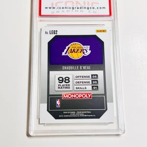 May include: A basketball trading card featuring Shaquille O'Neal in a protective case. The card displays the Los Angeles Lakers logo, player stats, and the text "MONOPOLY". The top of the case has the website address "www.iconicgradingco.com".