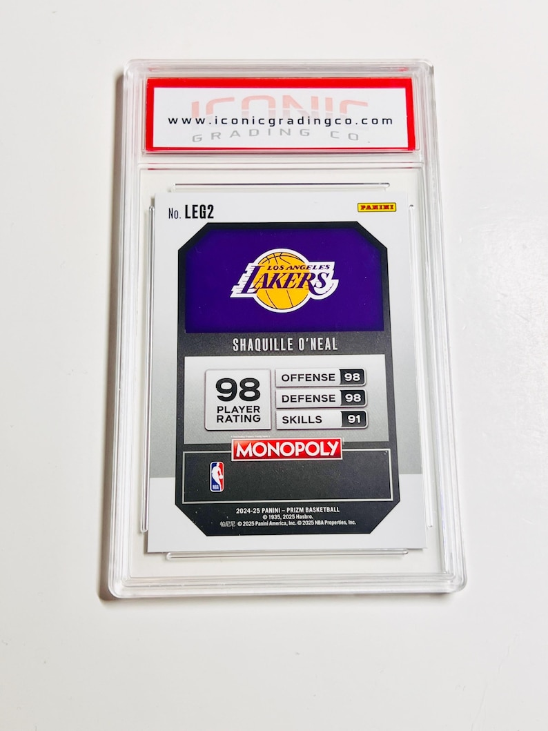May include: A basketball trading card featuring Shaquille O'Neal in a protective case. The card displays the Los Angeles Lakers logo, player stats, and the text "MONOPOLY". The top of the case has the website address "www.iconicgradingco.com".