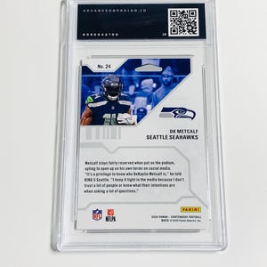 K&ouml;nnte beinhalten: Eine bewertete DK Metcalf Seattle Seahawks Footballkarte in einer Schutzh&uuml;lle. Die Karte zeigt ein Foto des Spielers in seiner Uniform. Die Karte ist mit der Nummer 24 versehen und enth&auml;lt Text &uuml;ber den Spieler.