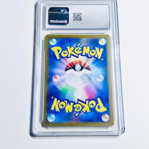 Op de afbeelding: Een beoordeelde Pok&eacute;mon-verzamelkaart in een doorzichtige plastic houder. De achterkant van de kaart is blauw met het Pok&eacute;mon-logo en een Pok&eacute; Ball-ontwerp. De houder heeft een label met de tekst "ggcards.com/verify".