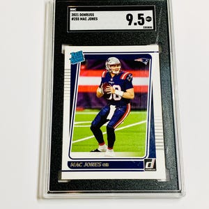 May include: A graded 2021 Donruss Mac Jones football card. The card shows the player in a blue and red uniform. The card is in a black holder, graded 9.5 by SOC. The text on the card reads "MAC JONES QB".