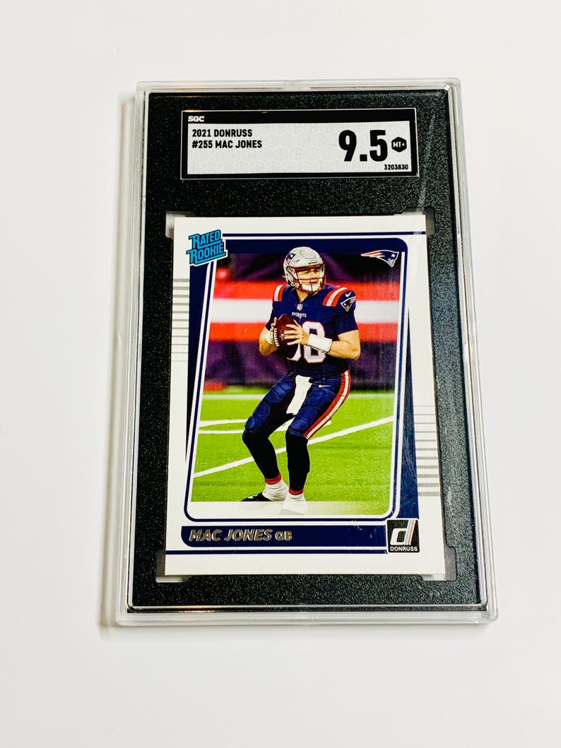 May include: A graded 2021 Donruss Mac Jones football card. The card shows the player in a blue and red uniform. The card is in a black holder, graded 9.5 by SOC. The text on the card reads "MAC JONES QB".