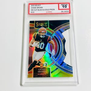 May include: A Chase Brown football card from the 2023 Select series, featuring a die-cut black and gold Prizm design. The card is graded Pristine 10 and includes the player's name and team, the Bengals. The card is encased in a protective holder.