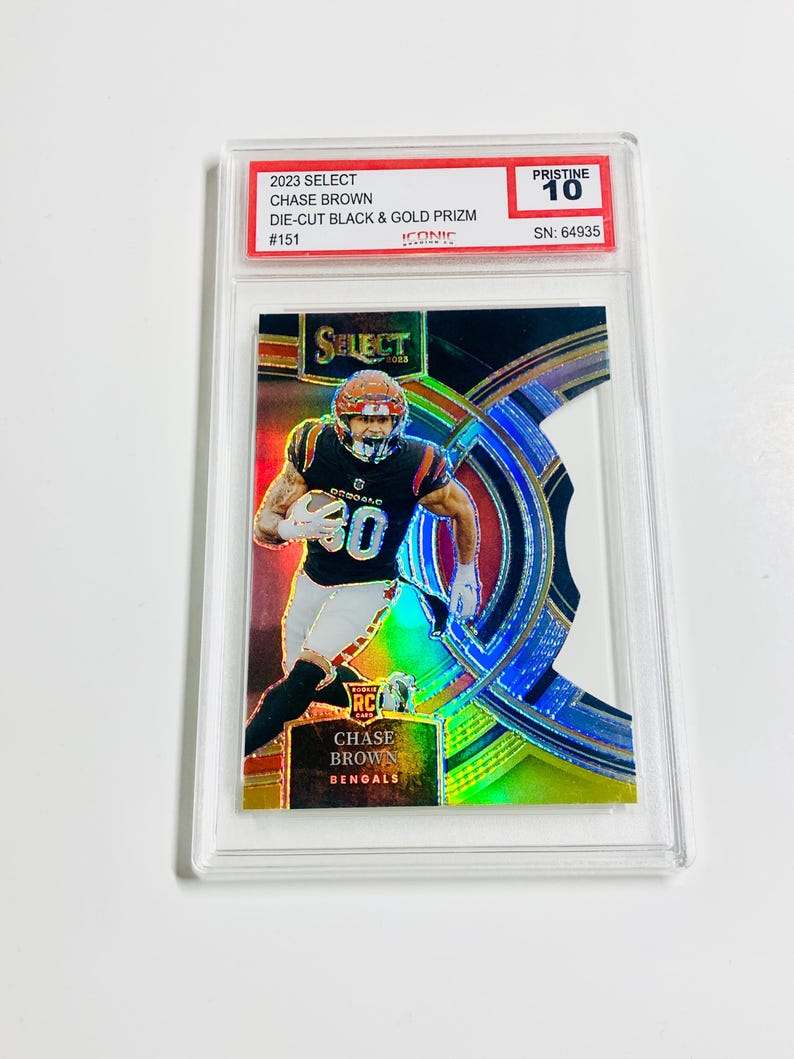 May include: A Chase Brown football card from the 2023 Select series, featuring a die-cut black and gold Prizm design. The card is graded Pristine 10 and includes the player's name and team, the Bengals. The card is encased in a protective holder.