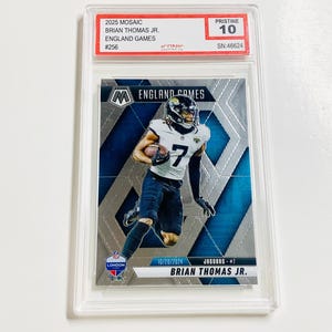 May include: A football trading card featuring Brian Thomas Jr. in a Jacksonville Jaguars uniform. The card is encased in a protective holder and includes the text "2025 Mosaic England Games" and "Pristine 10".