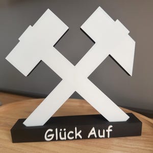 May include: A white and black decorative sculpture featuring crossed hammers, with the German phrase "Glück Auf" at the base. The sculpture is made of a solid material and is displayed on a wooden surface.