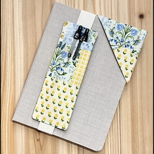 Puede incluir: Un cuaderno gris con un portalápices de tela y un marcapáginas a juego. El portalápices y el marcapáginas presentan un diseño de patchwork con estampados florales en azul, amarillo y blanco. Dos bolígrafos negros están en el portalápices.