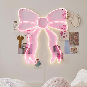 May include: A pink neon bow-shaped wall decor with a white outline. The bow is mounted on a white wall with photos and a key. The words "GOOF VIBE ONLY" are visible on a small card. The decor has a power cord.