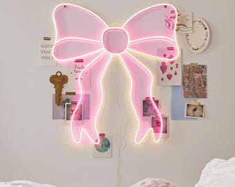 Pode incluir: Uma decoração de parede em forma de laço de néon rosa com um contorno branco. O laço é montado numa parede branca com fotos e uma chave. As palavras "GOOF VIBE ONLY" são visíveis num pequeno cartão. A decoração tem um cabo de alimentação.