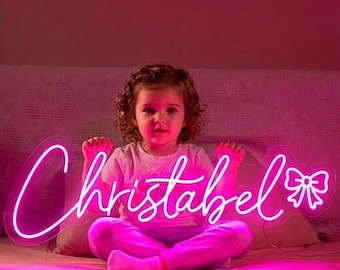 May include: A neon sign in a cursive font spelling "Christabel" with a bow at the end, illuminated in bright pink. The sign is held by a young child, seated on a bed, wearing a light pink shirt and pants.