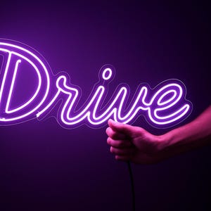 May include: A neon sign in the shape of the word "Drive" glows brightly in a vibrant purple hue. The sign is held by a person's hand against a dark background, creating a striking contrast.