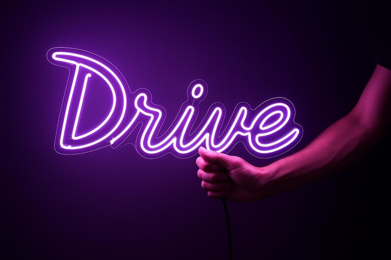 May include: A neon sign in the shape of the word "Drive" glows brightly in a vibrant purple hue. The sign is held by a person's hand against a dark background, creating a striking contrast.