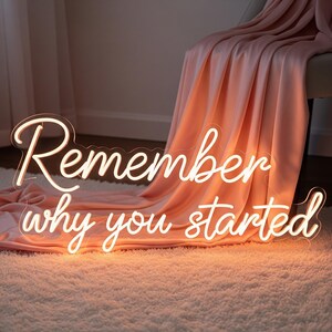 以下が含まれることがあります： 「Remember why you started」という言葉が筆記体で書かれたネオンサイン。温かみのある白色に光り、ふわふわの白いラグの上に置かれています。背景には、桃色の毛布が椅子にかけられています。