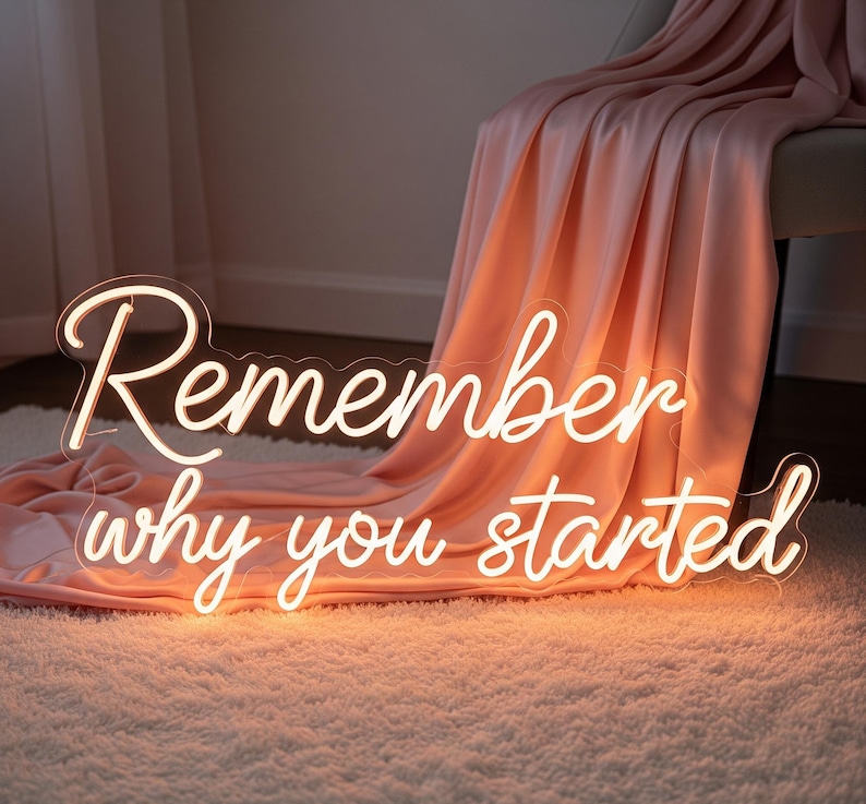 以下が含まれることがあります： 「Remember why you started」という言葉が筆記体で書かれたネオンサイン。温かみのある白色に光り、ふわふわの白いラグの上に置かれています。背景には、桃色の毛布が椅子にかけられています。