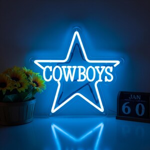 以下が含まれることがあります： 白い「COWBOYS」の文字が入った、星型の青いネオンサイン。暗い背景に光っています。小さな木製のカレンダーには「JAN 60」と表示され、ひまわりのバスケットの隣にあります。
