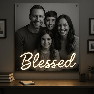 May include: A black and white family portrait with the word "Blessed" in neon script. The portrait is mounted on a wall, with framed pictures and a desk with books and a plant in front.