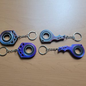 May include: Four keychains with a bearing design. The keychains have a metallic finish with shades of blue, purple, and red. Each keychain has a silver chain and a key ring. The keychains are on a light brown wooden surface.