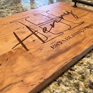 May include: A rectangular wooden serving tray with a light brown finish. The tray features the name "Henry" and the text "ESTABLISHED 2023" engraved on the surface. It has two black metal handles.