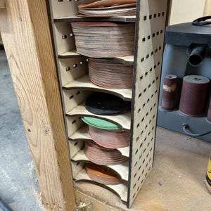 May include: A wooden storage rack with six shelves for holding sanding discs. The rack is made of plywood and has a light brown finish. The sanding discs are stacked on each shelf and are of various colors, including red, brown, green, and black.