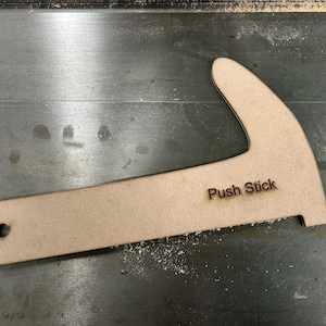 Könnte beinhalten: Ein hellbrauner Holz-Schiebestock mit dem eingebrannten Text "Push Stick". Das Werkzeug hat einen gebogenen Griff und eine rechteckige Basis mit einem Loch. Für die Holzbearbeitung.