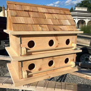May include: A two-tier wooden birdhouse with a shingled roof. Each level features three circular entry holes and a wooden dowel perch. The birdhouse is constructed from light-coloured wood and is designed for outdoor use.