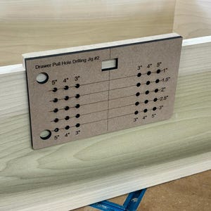 Op de afbeelding: Een bruine houten lade-trekgat-boorjig met een zwarte rand. De jig heeft een raster van gaten met metingen in inches voor het boren van lade-trekgaten. De tekst "Drawer Pull Hole Drilling Jig #2" is op de jig gedrukt.