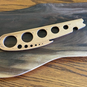 May include: A light-colored wooden ring sizer with multiple circular openings of varying sizes. The tool is labeled with Roman numerals and the word "Child". The text "Golden Grove Creations" is also visible. The sizer is resting on a dark wooden cutting board.