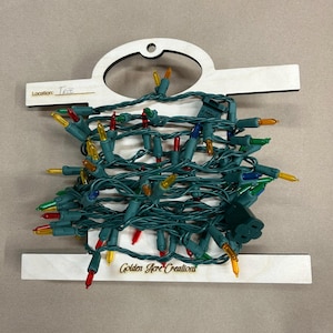 May include: A bundle of multi-colored Christmas lights on a wooden holder. The lights are green with red, yellow, blue, and green bulbs. The holder has the text "Location: Tree" and "Golden Hare Creations".