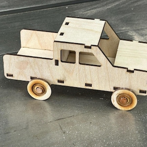May include: A wooden toy pickup truck with a natural wood finish. The truck features a detailed design with a cab, bed, and four wooden wheels. The wheels have a circular design with a darker wood center.