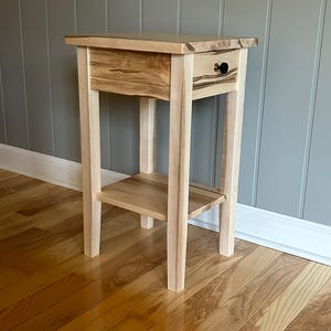 May include: A light-colored wooden side table with a drawer and a lower shelf. The table has four legs and a dark knob on the drawer. The wood grain is visible on the top and drawer front. The table is approximately 28 inches tall.