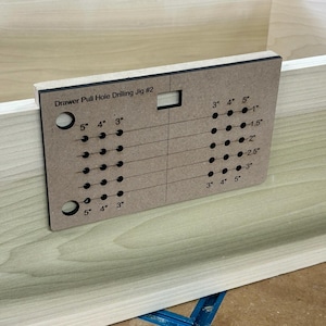 May include: A brown wooden drawer pull hole drilling jig with a black border. The jig has a grid of holes with measurements in inches for drilling drawer pull holes. The text "Drawer Pull Hole Drilling Jig #2" is printed on the jig.