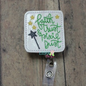 May include: A white square badge holder with glitter and green and gold embroidery that reads "Faith Trust Pixie Dust". A black wand with a green star is embroidered on the left side of the badge. The badge is attached to a clear plastic retractable badge reel.