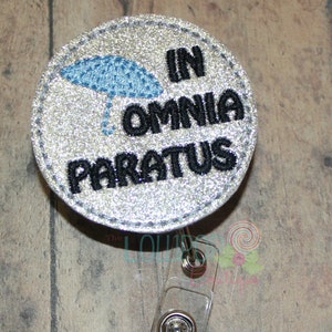 May include: Silver glitter badge with a blue umbrella and the text "In Omnia Paratus" in black embroidery. The badge is attached to a clear plastic retractable badge holder.