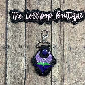 May include: Purple and green embroidered flower keychain with a black leather backing and a silver metal clasp. The keychain is attached to a black background.