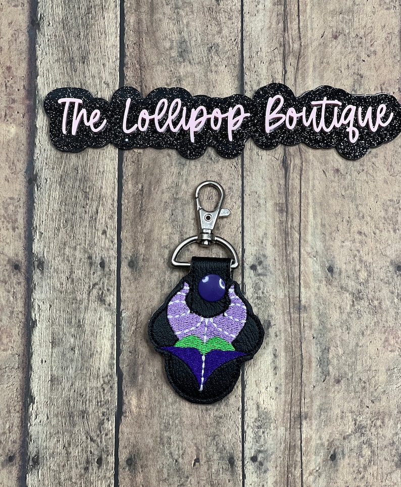 May include: Purple and green embroidered flower keychain with a black leather backing and a silver metal clasp. The keychain is attached to a black background.