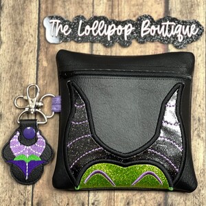 May include: Black zippered pouch with a green and purple embroidered design of Maleficent's horns and face. The pouch is attached to a silver key chain with a purple and green embroidered flower charm. The text "The Lollipop Boutique" is visible on a black and silver banner.