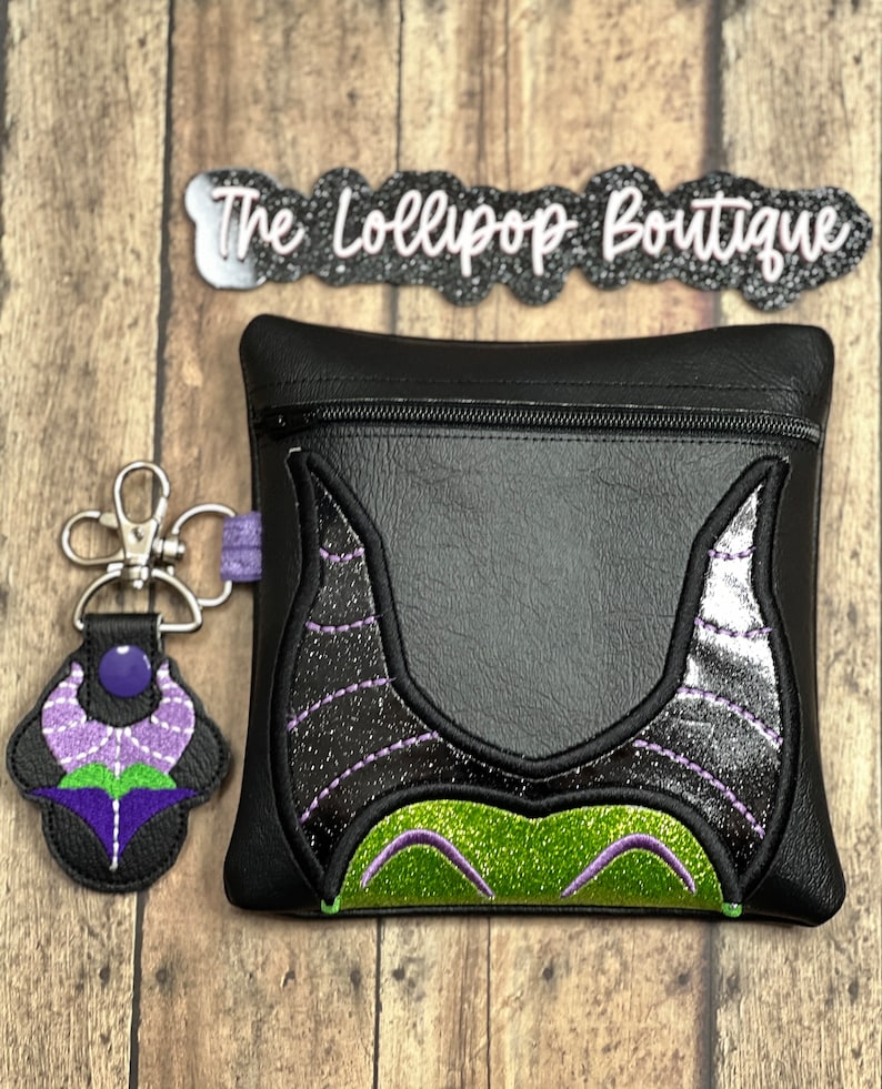 May include: Black zippered pouch with a green and purple embroidered design of Maleficent's horns and face. The pouch is attached to a silver key chain with a purple and green embroidered flower charm. The text "The Lollipop Boutique" is visible on a black and silver banner.