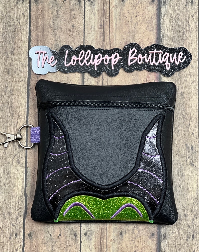 May include: Black zippered pouch with a green and black glitter Maleficent design. The pouch has a purple zipper pull and a silver key ring.